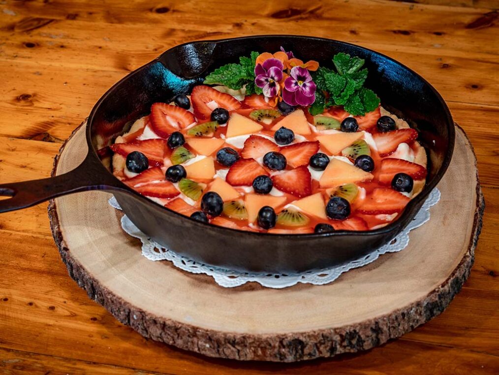 Cast-iron skillet fruit tart topped with sliced strawberries, kiwi, peaches, and blueberries, garnished with edible flowers on a wooden board.
