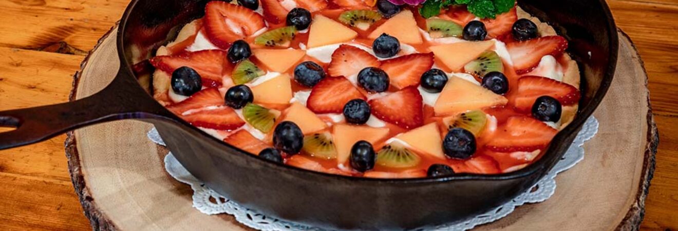 Cast-iron skillet fruit tart topped with sliced strawberries, kiwi, peaches, and blueberries, garnished with edible flowers on a wooden board.